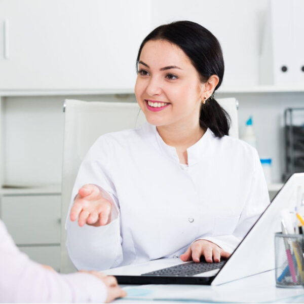 Lakeshore Bone & Joint Orthopedic Urgent Care | A female doctor in a white coat is sitting at a desk with a laptop, talking to a patient. The doctor is smiling and gesturing with her hand.