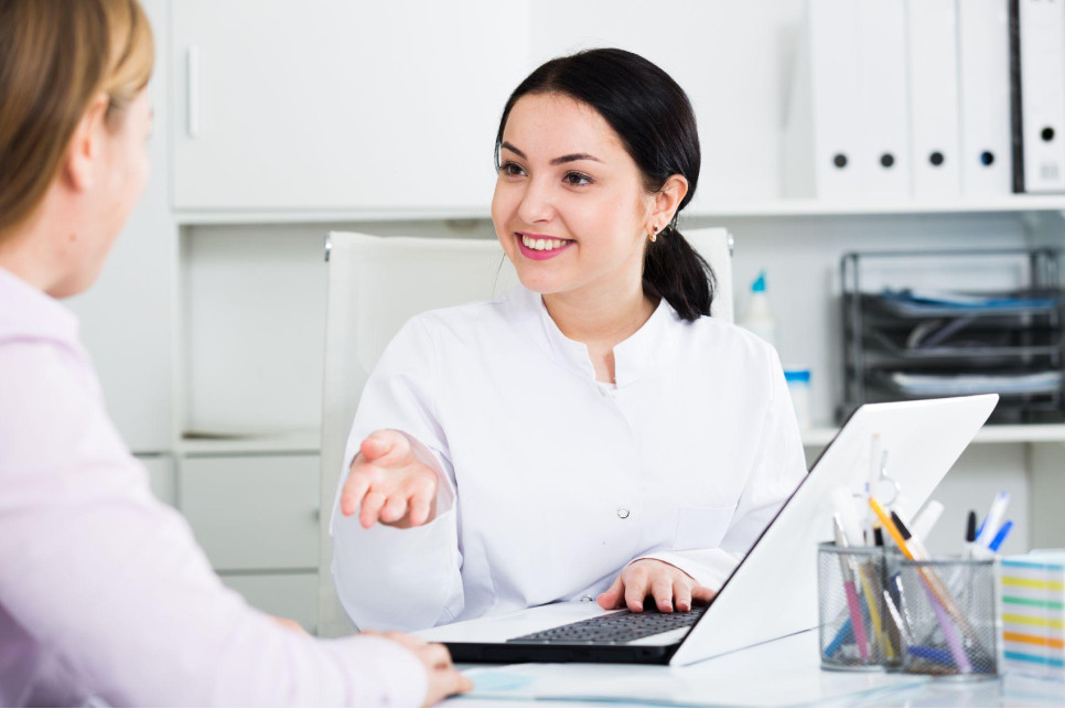 Lakeshore Bone & Joint Orthopedic Urgent Care | A female doctor in a white coat is sitting at a desk with a laptop, talking to a patient. The doctor is smiling and gesturing with her hand.