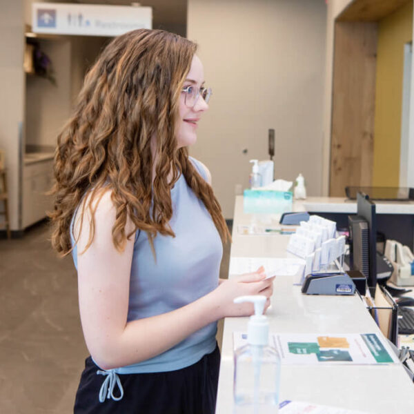 Lakeshore Bone & Joint Orthopedic Urgent Care | A young woman with glasses stands at a reception desk in what appears to be a medical or office setting, holding paperwork and wearing a light blue sleeveless top.